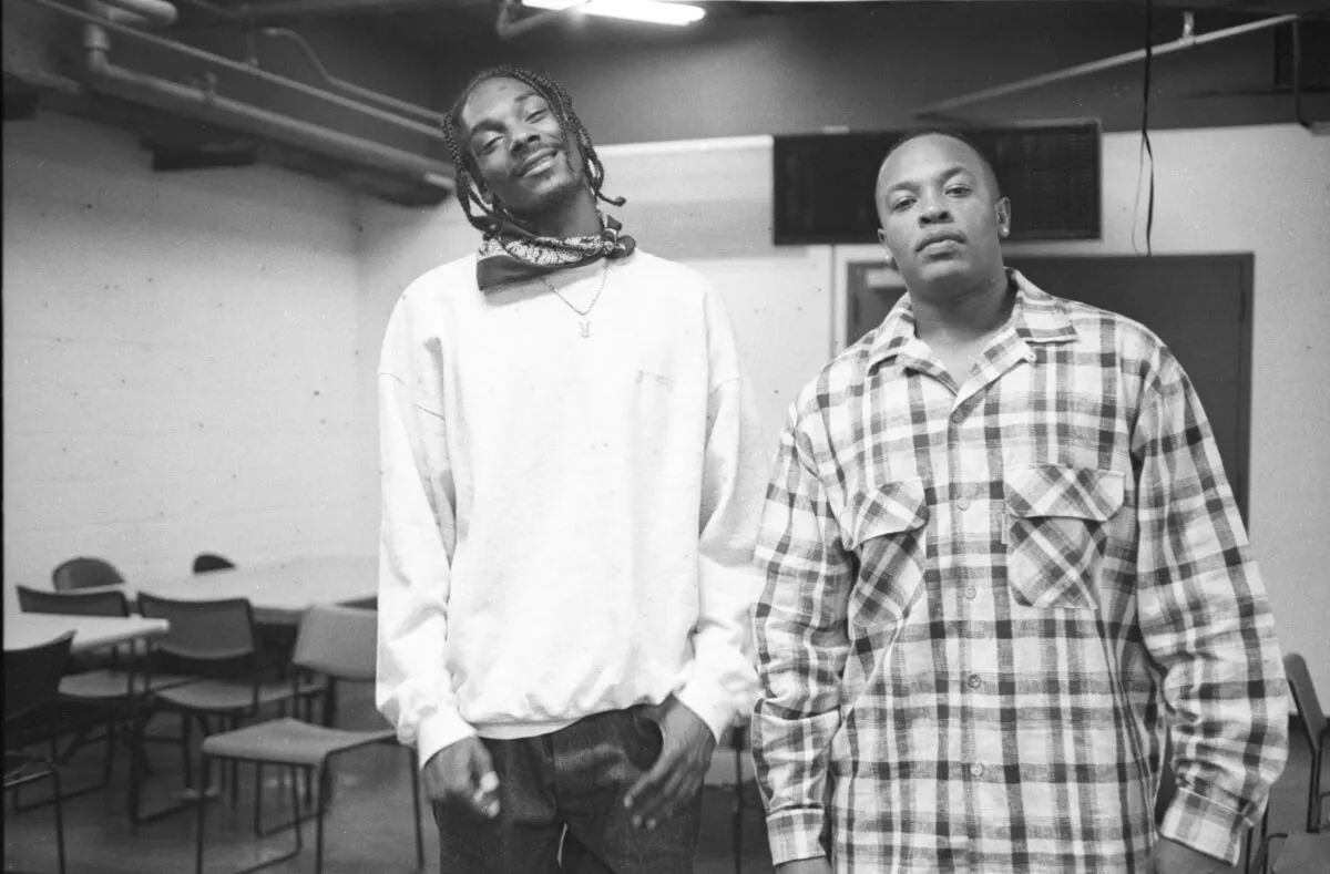 A black and white picture of Snoop Dogg and Dr. Dre standing together. Snoop wears a white shirt with a bandanna around his neck. Dr. Dre wears a flannel shirt.