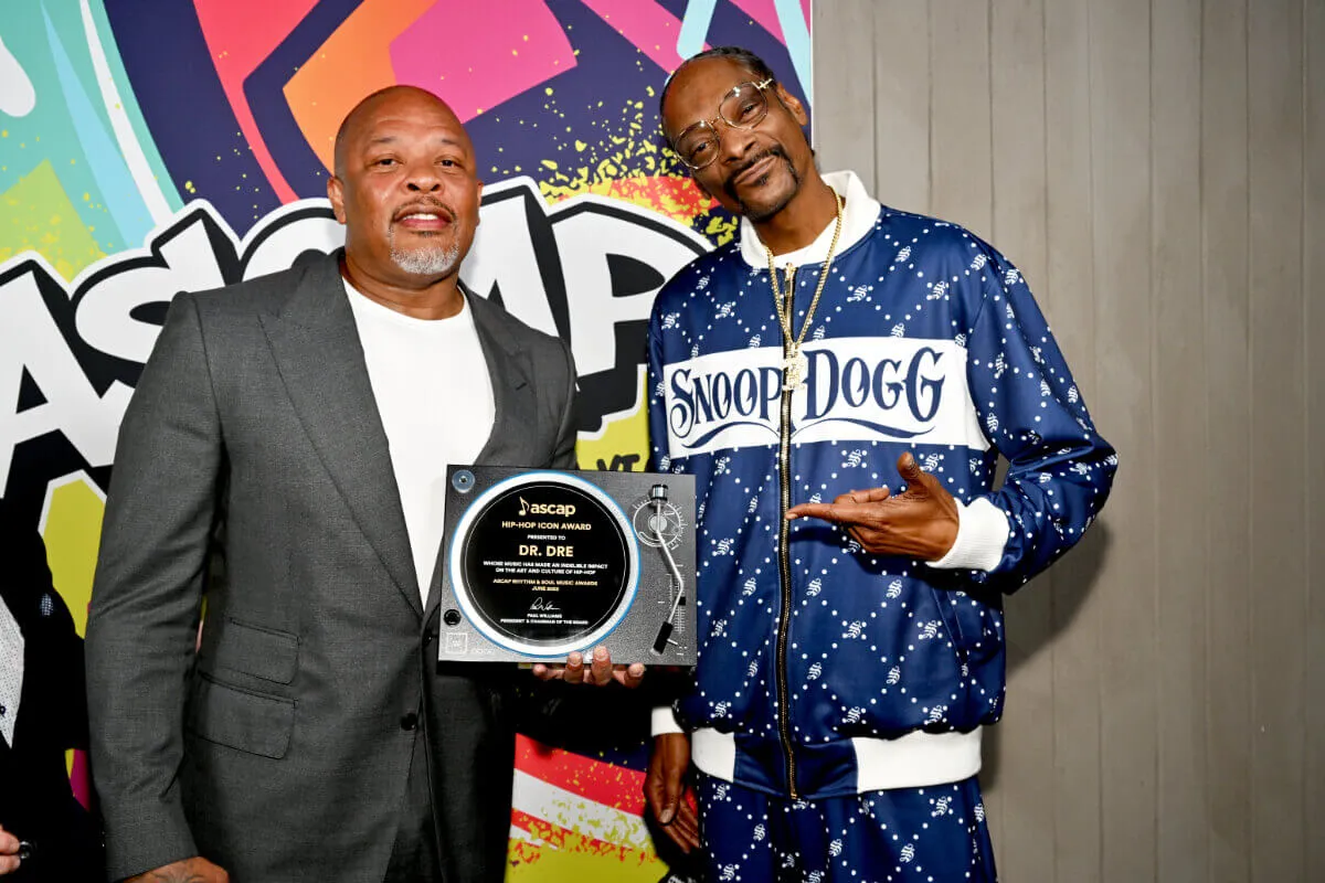 Dr. Dre wears a suit and stands with Snoop Dogg, who wears a blue personalized track suit. Dr. Dre holds an ASCAP award between them.