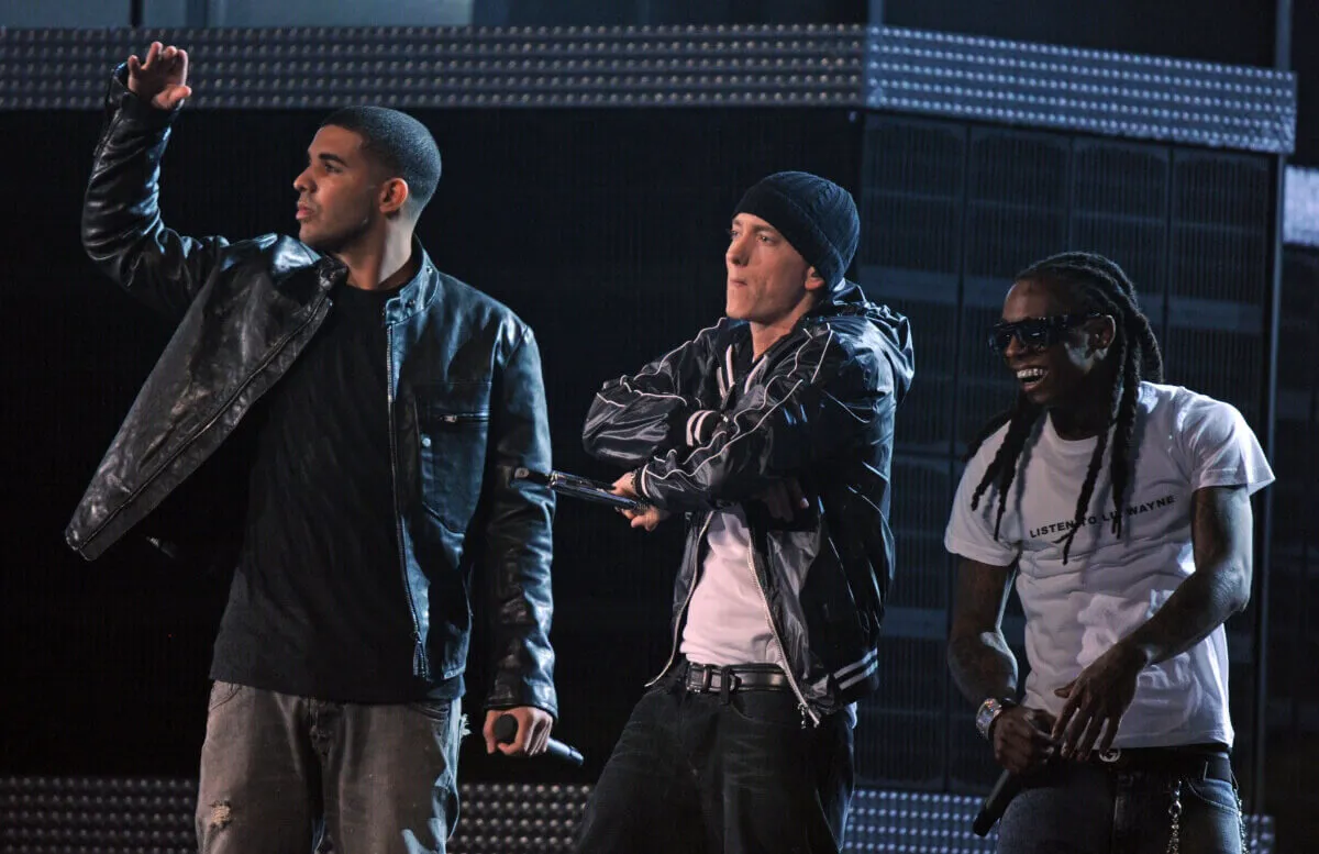 Drake, Eminem, and Lil Wayne stand together. Drake lifts his arm up. Drake and Eminem wear black leather jackets and Lil Wayne wears a white shirt.