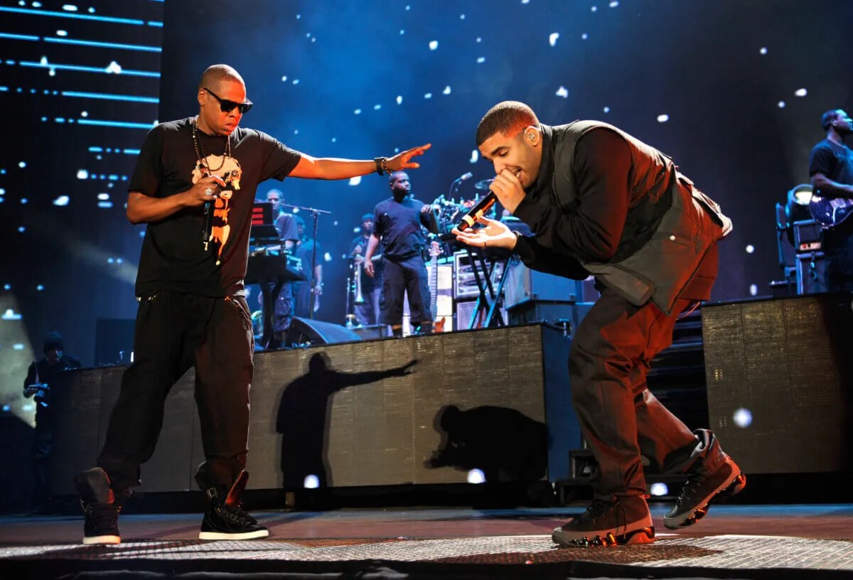 Jay-Z and Drake are on a stage together. Jay-Z wears sunglasses and holds a microphone. Drake bends towards the microphone.