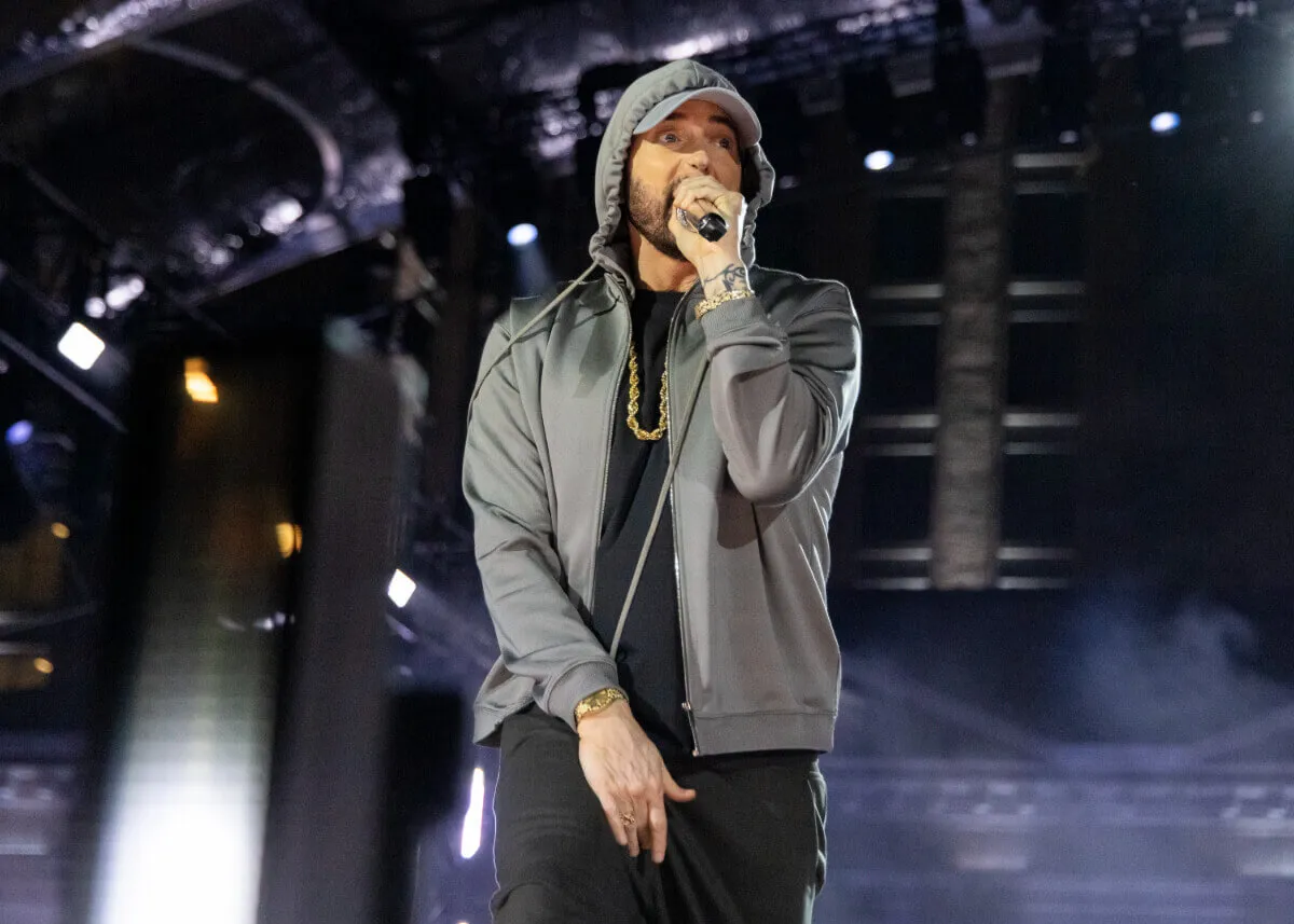 Eminem wears a gray hoodie and holds a microphone to his face.