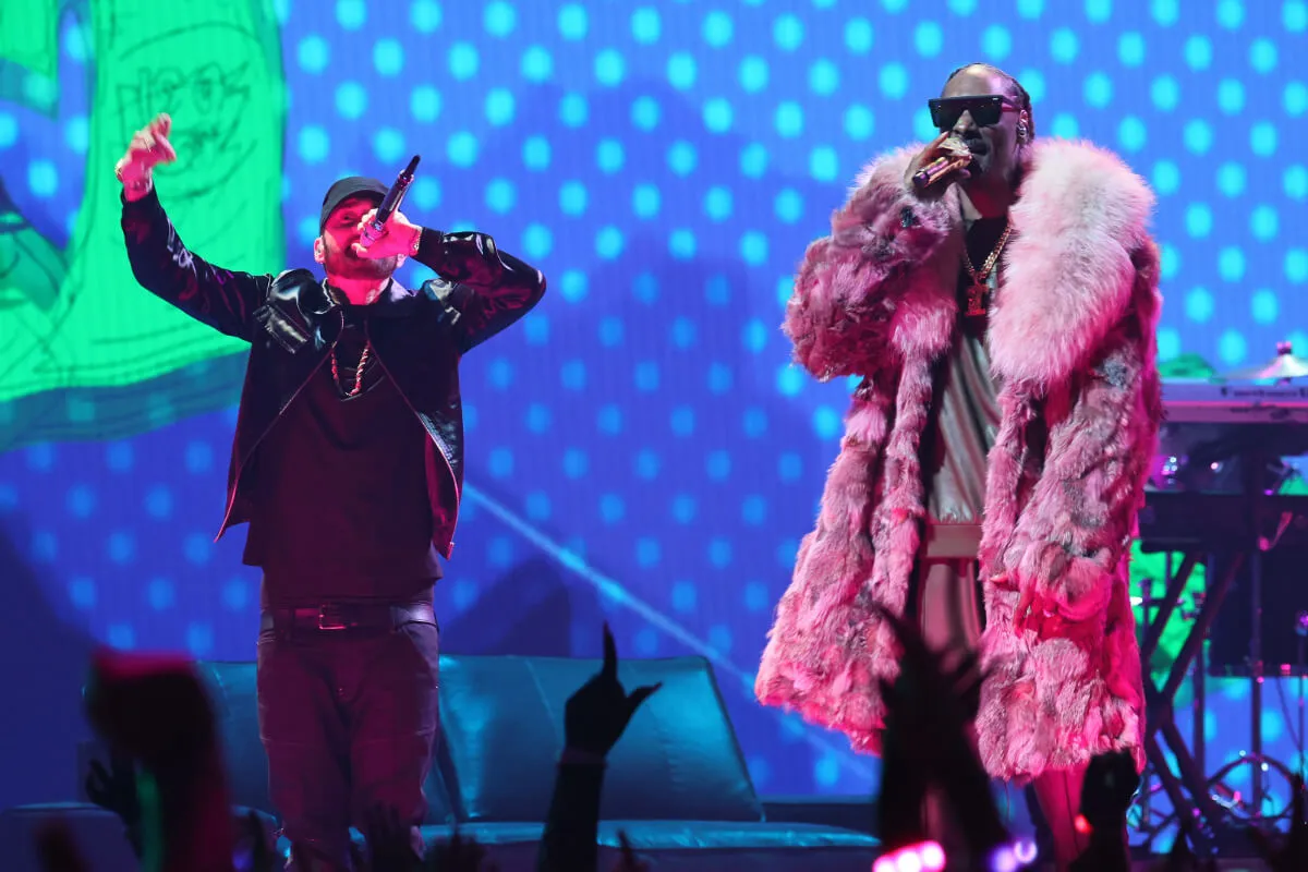 Eminem and Snoop Dogg stand on a stage together. Eminem holds his arms up and wears black. Snoop wears a coat and sunglasses.