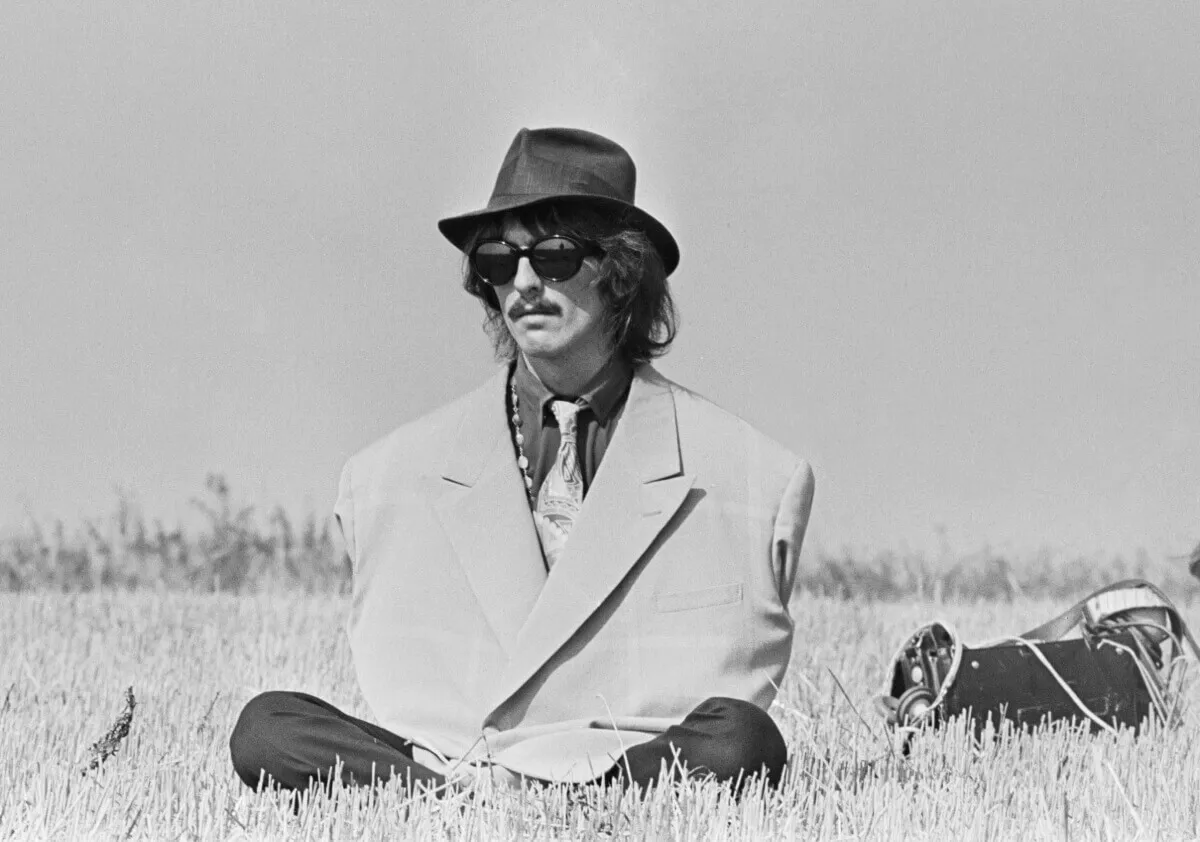 A black and white picture of George Harrison wearing an oversized blazer, sunglasses, and a hat. He sits cross legged in a field.