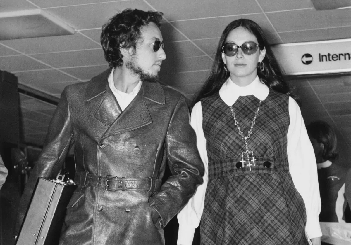 A black and white picture of Bob Dylan and Sara Lownds walking together. They both wear sunglasses.