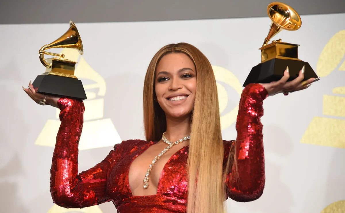 Beyonce holds up a Grammy award in each hand. She wears a red sparkly dress.