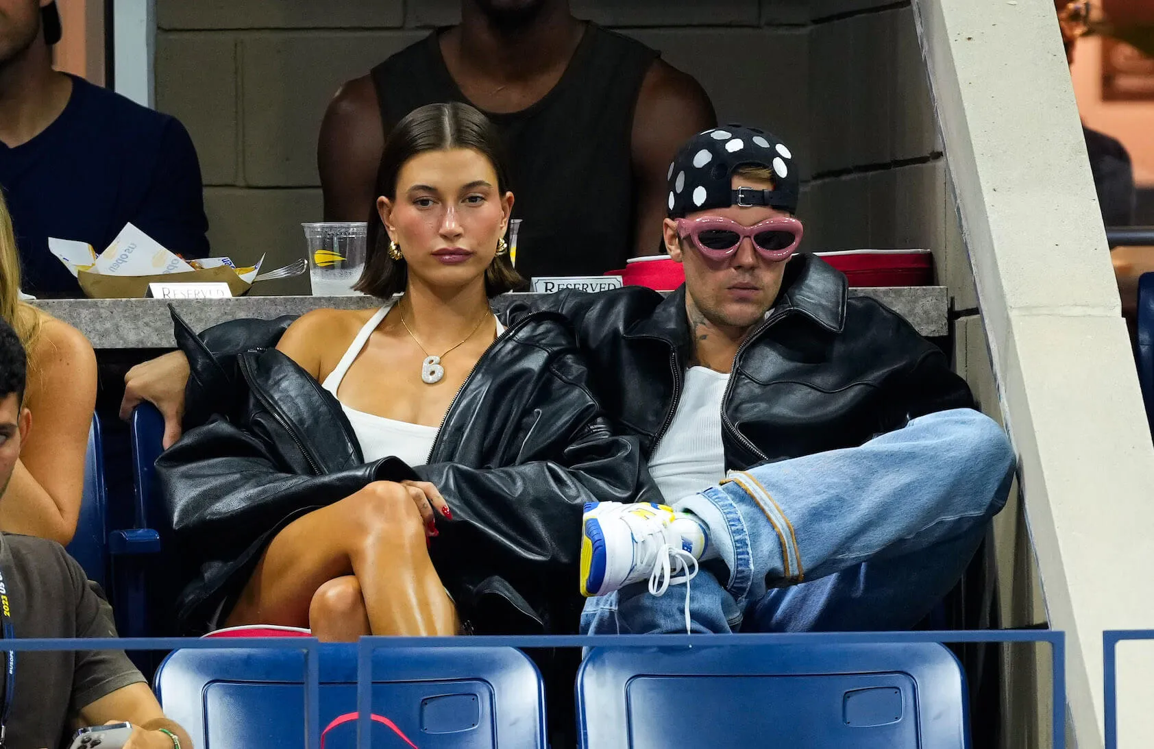 Hailey Bieber and Justin Bieber sitting next to each other at the US Open in 2023. Hailey is wearing a leather jacket falling off of her shoulders. Justin is wearing a leather jacket with pink sunglasses and a backward cap.