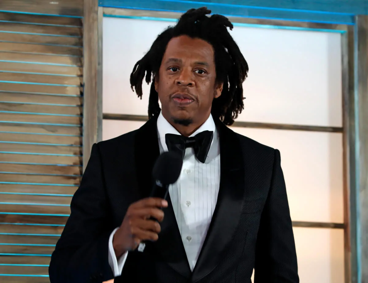 Jay-Z wears a tuxedo and holds a microphone.