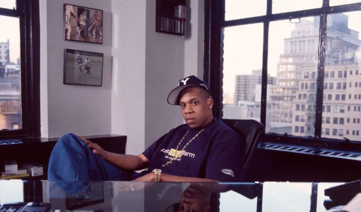 Jay-Z wears a black shirt and hat and sits at a desk.