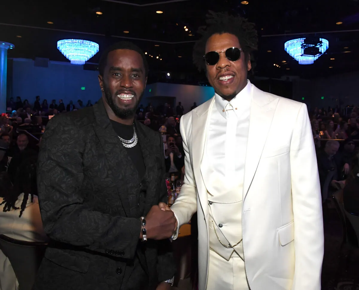 Sean 'Diddy' Combs wears black and shakes hands with Jay-Z who wears a white suit and sunglasses.