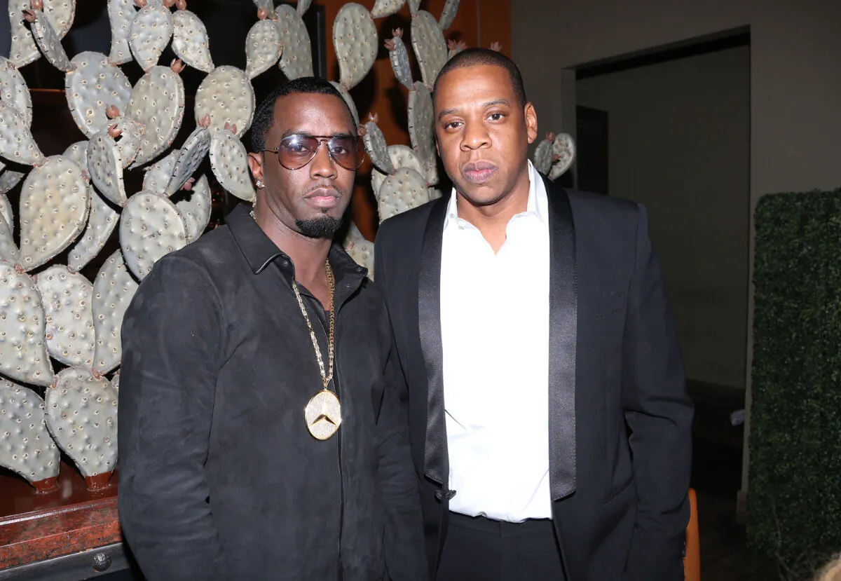 Sean 'Diddy' Combs and Jay-Z stand together. Combs wears sunglasses and a black shirt. Jay-Z wears a suit jacket.