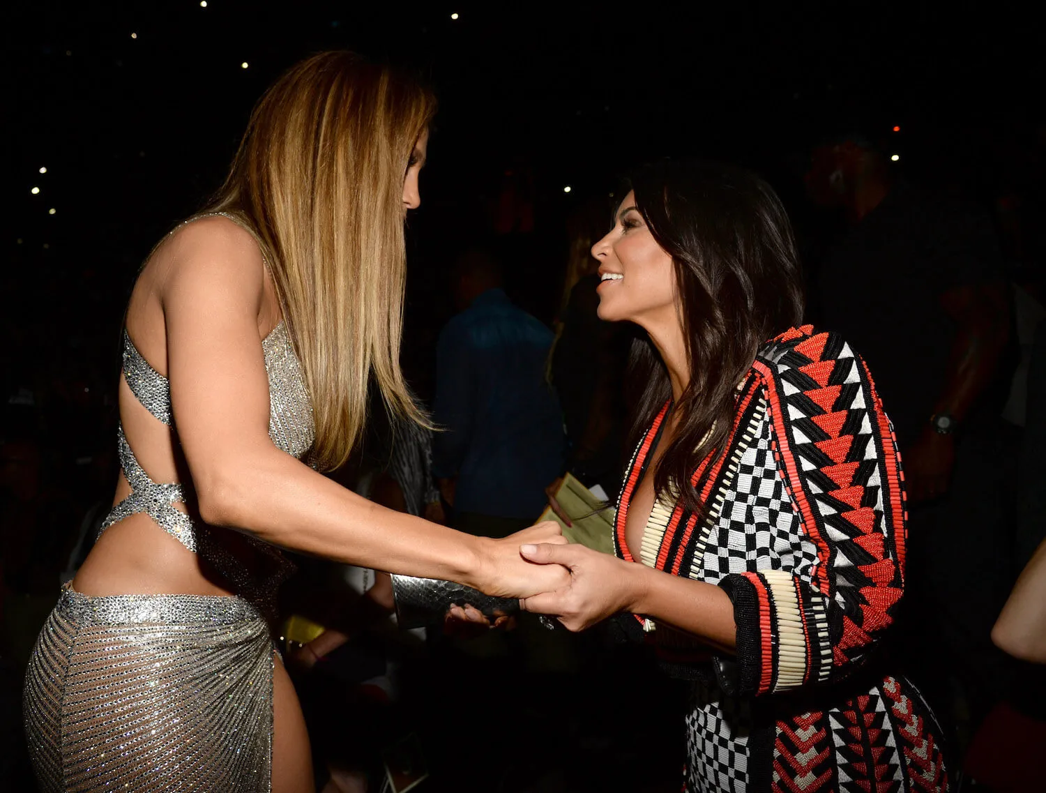 Jennifer Lopez and Kim Kardashian holding hands and facing each other while talking intensely against a black background