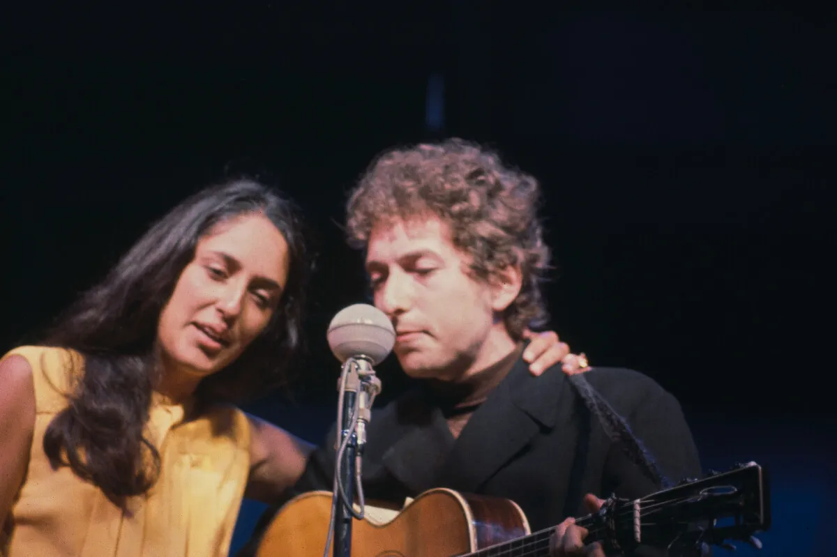 Joan Baez wears yellow and stands with her arm on Bob Dylan's shoulders. He wears black and holds a guitar.