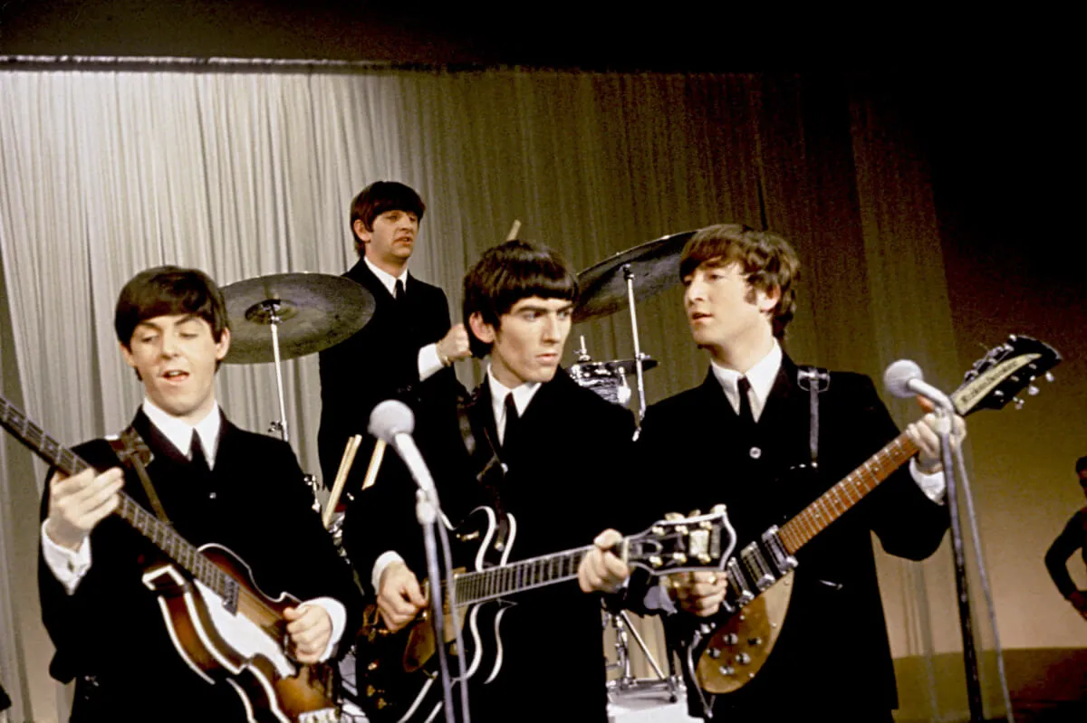 Paul McCartney, George Harrison, and John Lennon stand in front of microphones and play guitars. Ringo Starr plays drums on a platform behind them. They all wear suits.