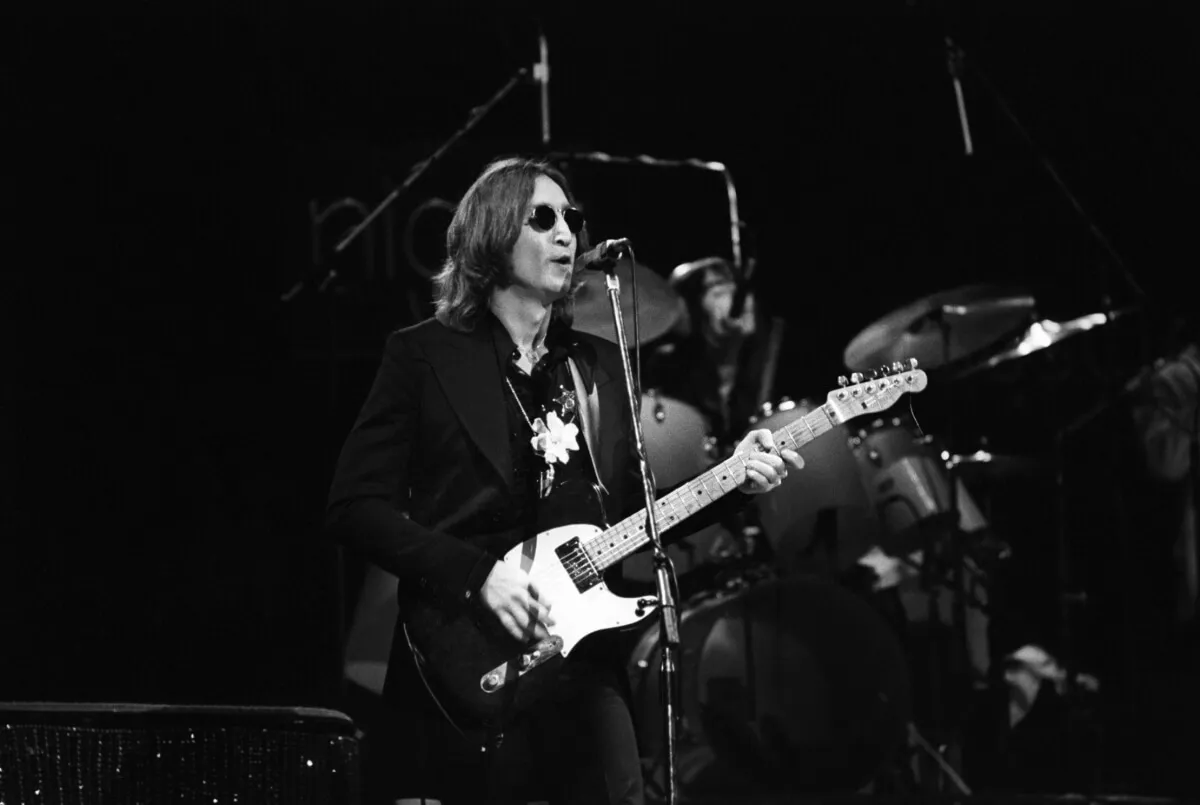 A black and white picture of John Lennon playing guitar and singing into a microphone. He stands on a stage and wears sunglasses.