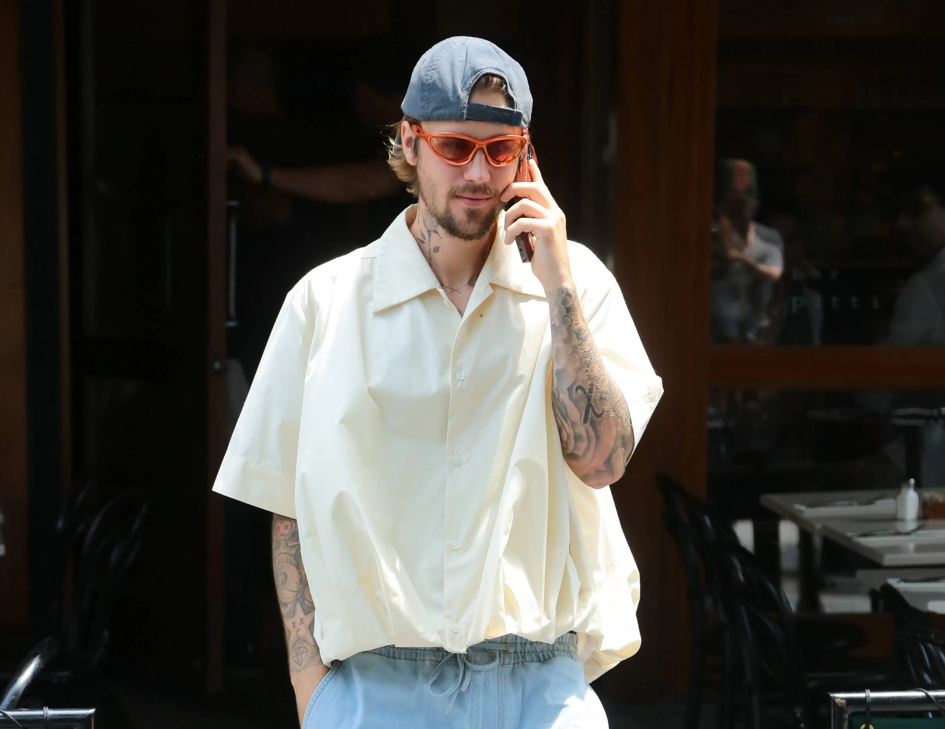 Justin Bieber talking on the phone during the day. He's wearing a loose-fitting short-sleeve button-up shirt with a backwards hat and orange sunglasses