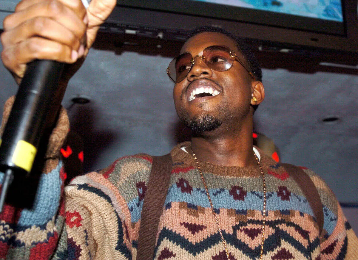 Kanye West wears a sweater and sunglasses. He holds a microphone.