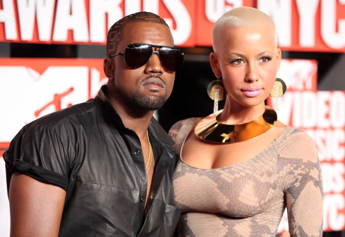 Kanye West wears a black shirt and sunglasses and stands with Amber Rose, who wears a gold necklace.