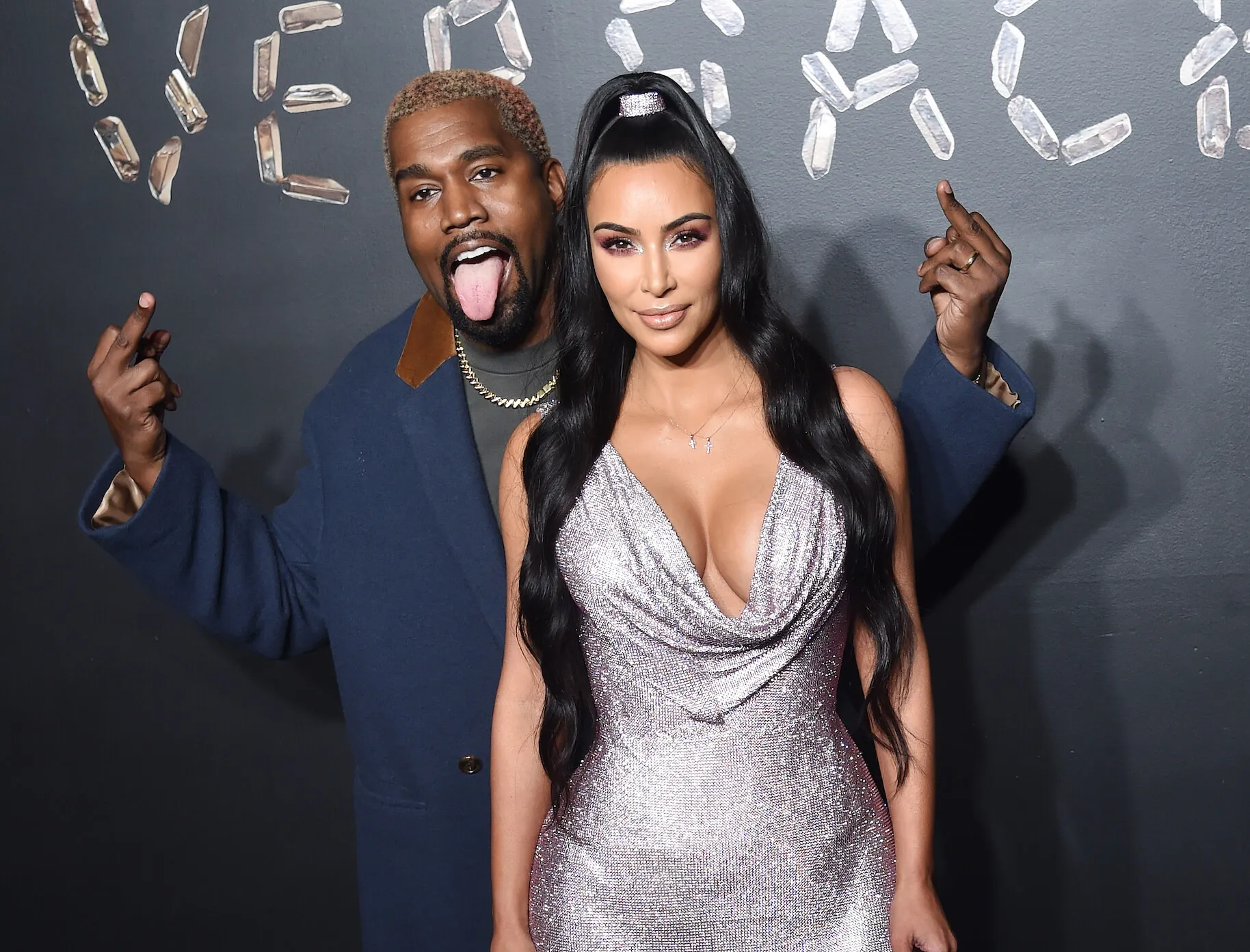 Kanye West standing behind Kim Kardashian with his hands up and his tongue out. Kardashian is smiling slightly and wearing a sparkling dress.