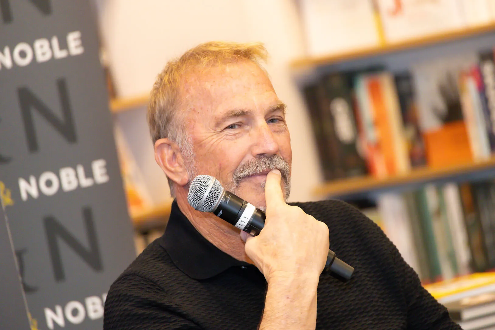 Kevin Costner holding a microphone and a finger up to his lips at a Barnes & Noble event