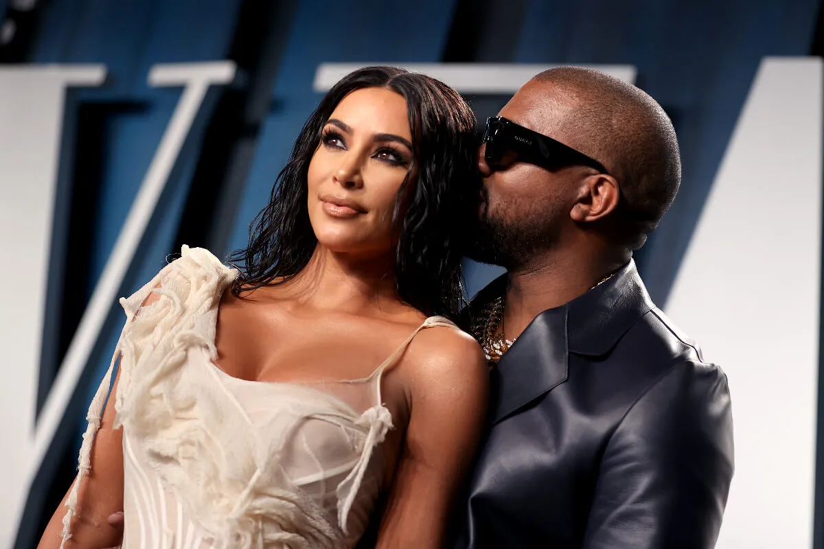 Kim Kardashian and Kanye West stand together. She wears a white dress and he wears a leather jacket and sunglasses. He's kissing the back of her head.