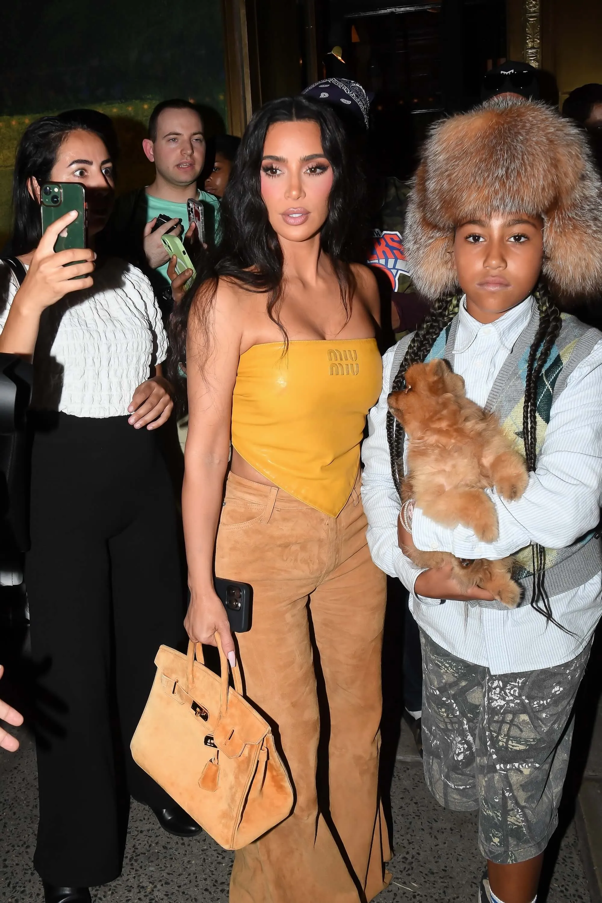 Kim Kardashian in a sleeveless yellow top and tan pants with North West in a fur hat that looks like Bianca Censori's 