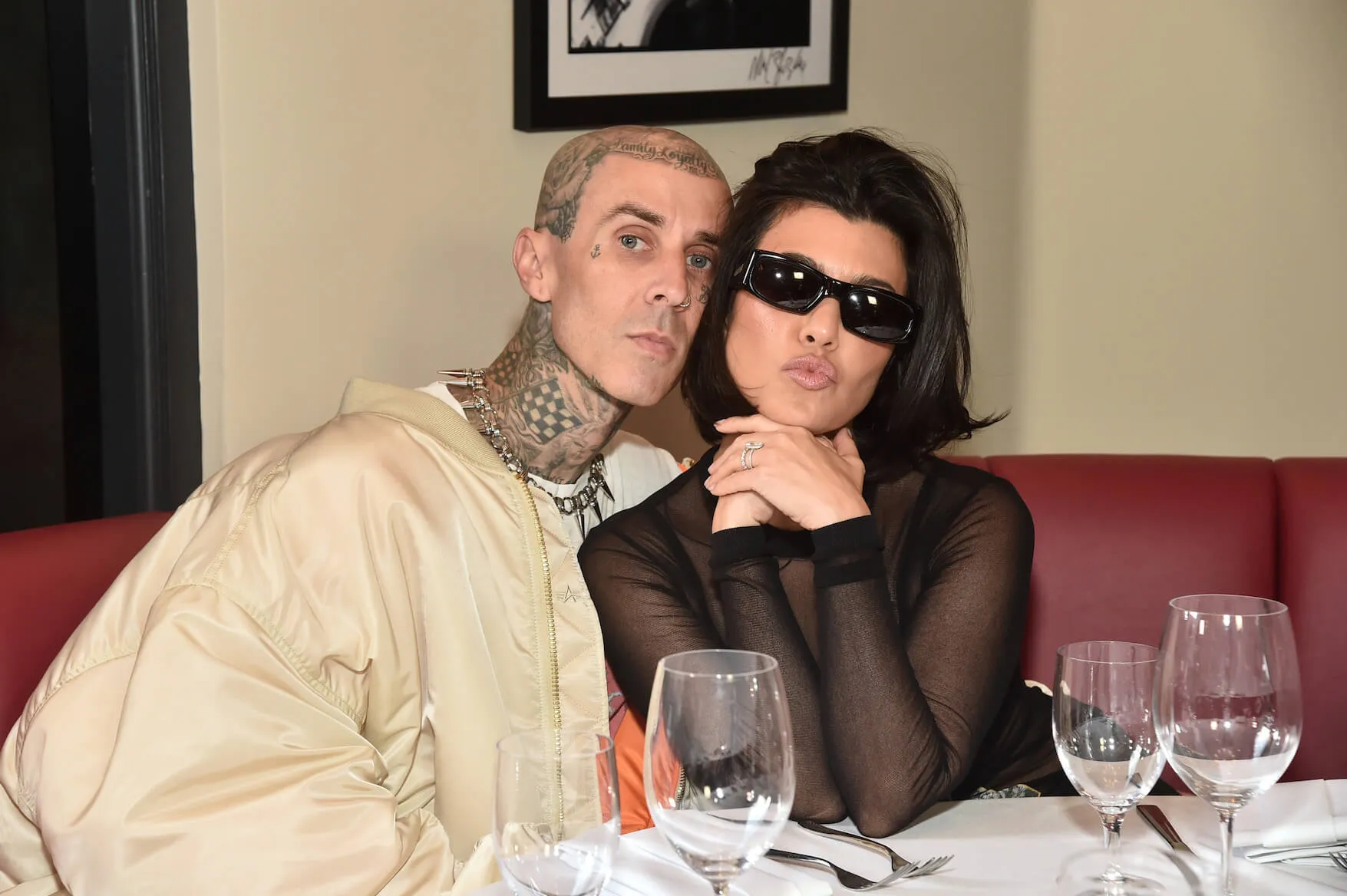 Travis Barker and Kourtney Kardashian sitting at a restaurant and posing for a photo. Kourtney has her lips pursed and sunglasses on.