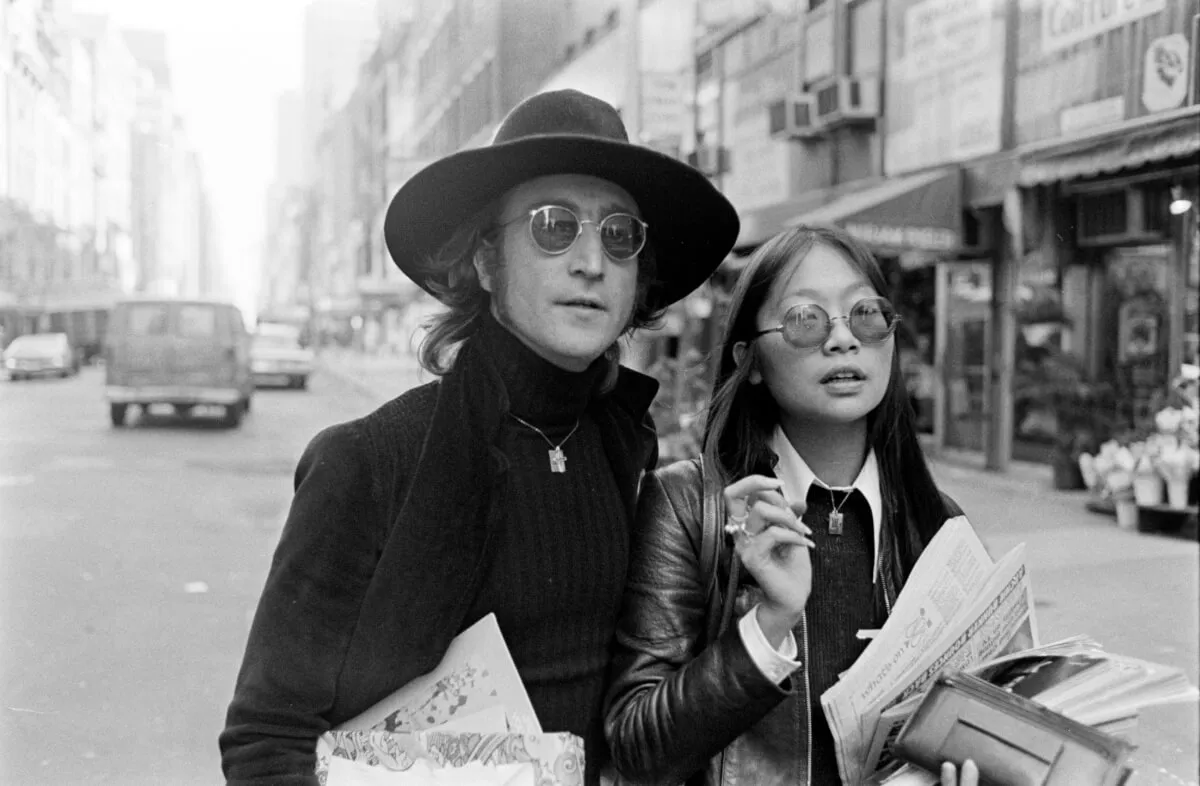 A black and white picture of John Lennon and May Pang walking in the street together. They both wear sunglasses and hold newspapers. Lennon wears a wide-brimmed hat.