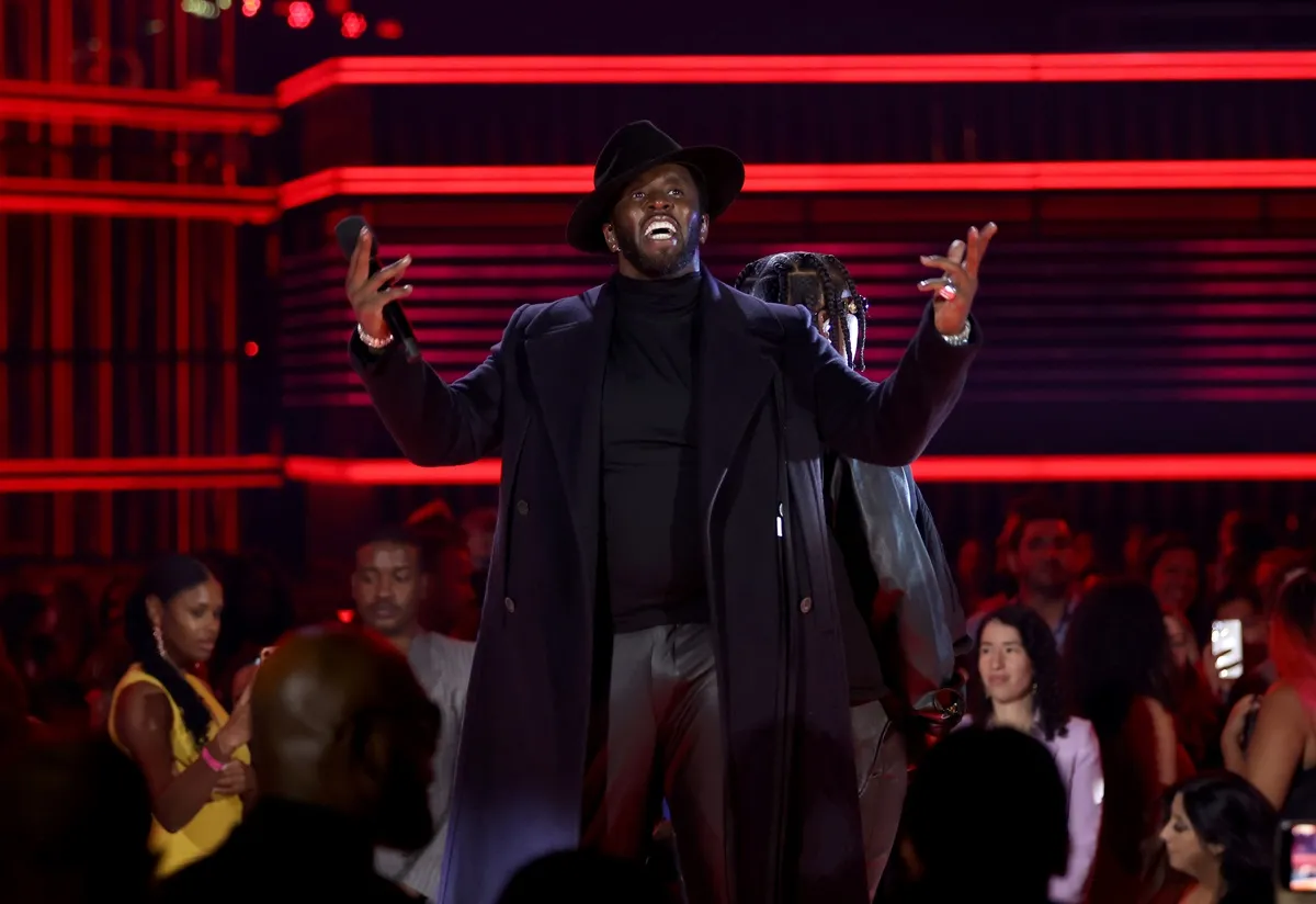 Sean Diddy Combs posing on stage while wearing a black trench coat, shirt, and hat.