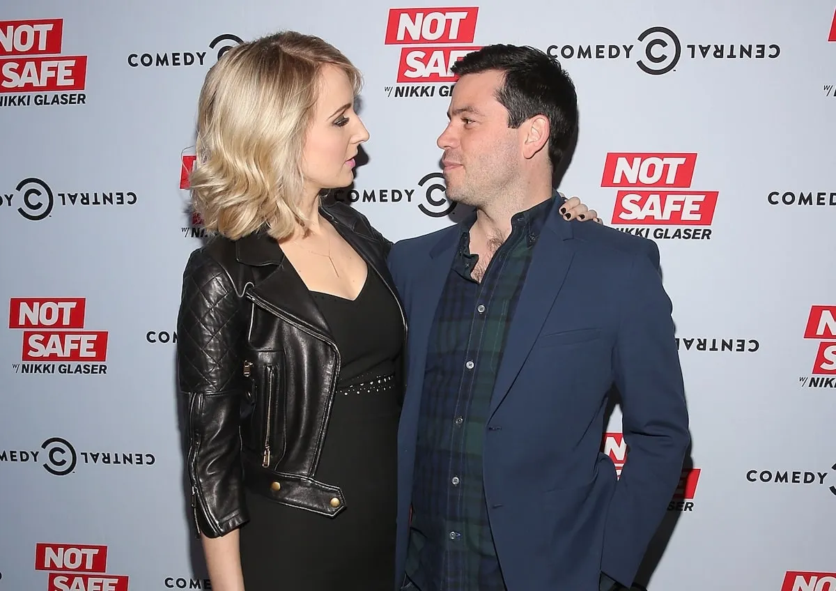 Wearing a black leather jacket and dress, Nikki Glaser smiles at her boyfriend and executive producer Chris Convy on a red carpet