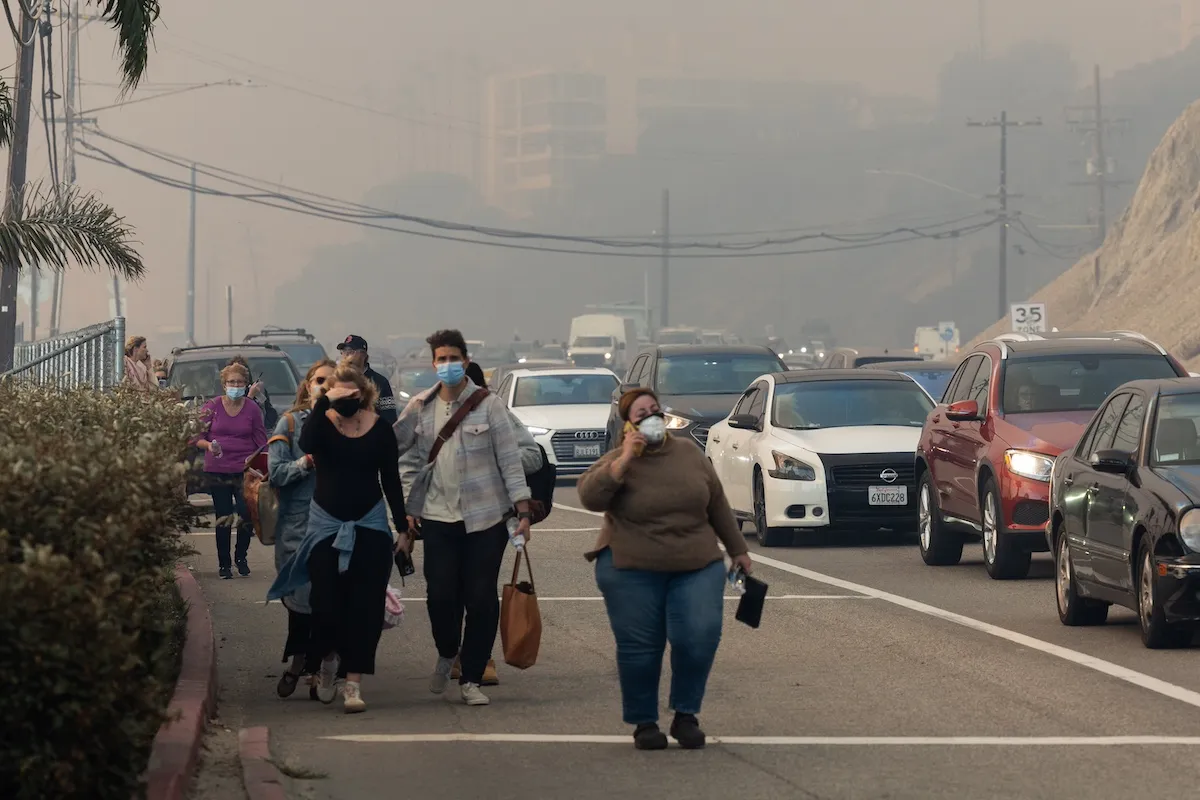 People fleeing a wildfire walk down the street in Pacific Palisades as car sit in a traffic jam