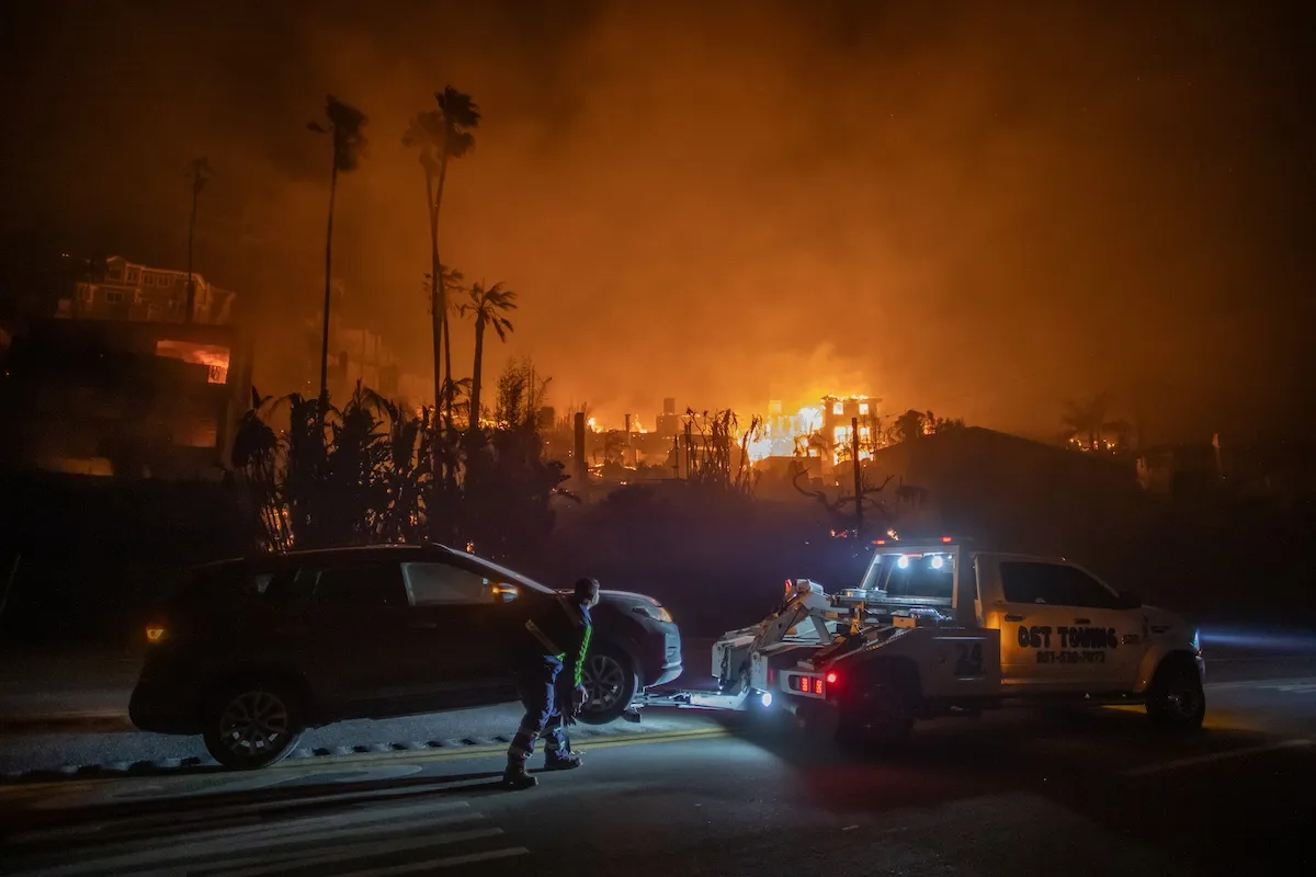 A tow truck towing a car at night as a wildfire rages in the background