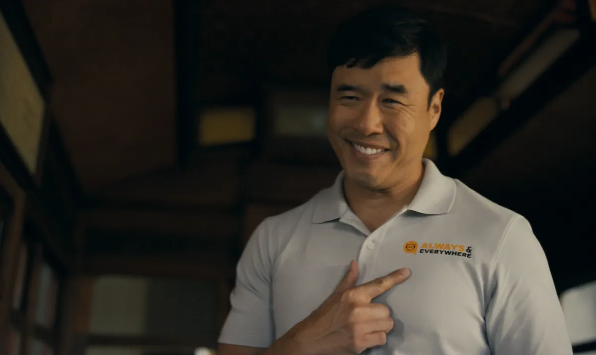 Randall Park pointing to the logo on his white polo shirt in 'Watson'