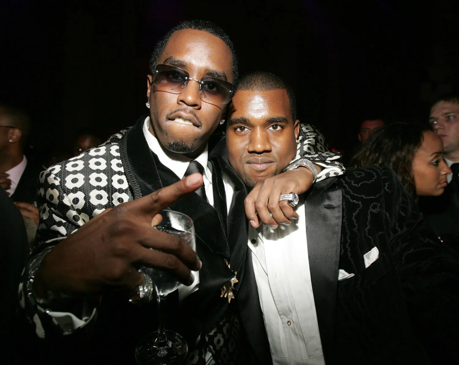 Sean 'Diddy' Combs wears a patterned jacket and sunglasses and stands with his arm around Kanye West's neck.