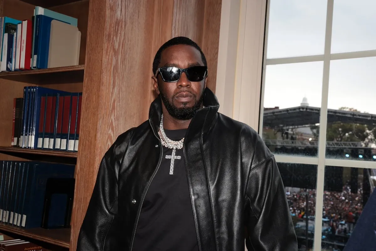 Sean 'Diddy' Combs wears a black jacket and sunglasses. He stands between a bookshelf and a window.