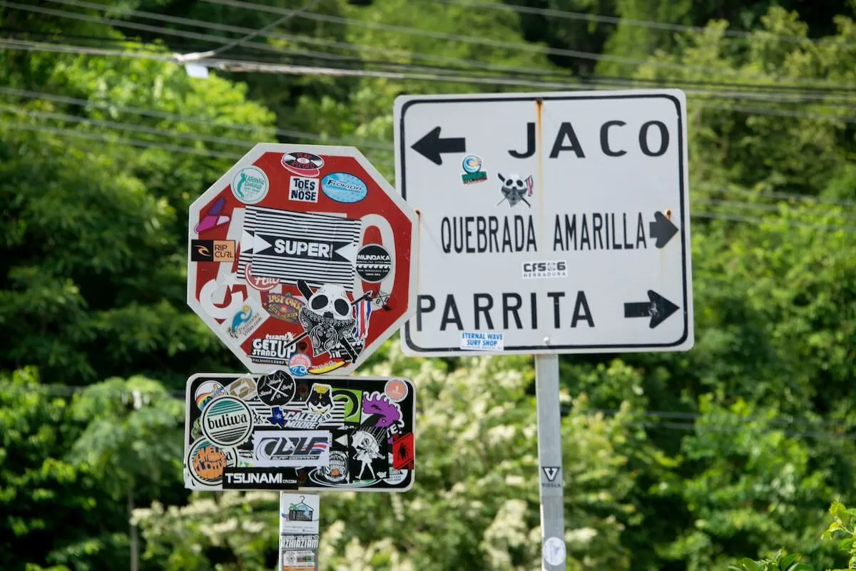 Road sign with arrows pointing to Jaco and Parrita next to a Stop sign covered with stickers