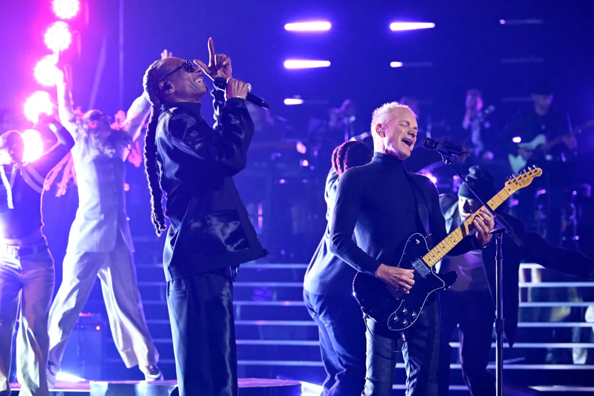 Snoop Dogg sings into a microphone and stands onstage with Sting. Sting plays guitar and stands in front of a microphone. They both wear black.