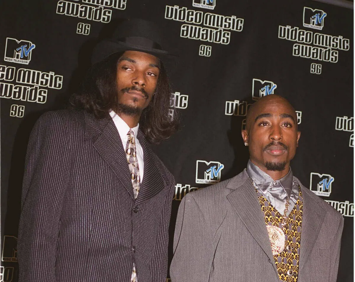 Snoop Dogg wears a suit and hat and stands next to Tupac, who wears a suit and yellow-patterned vest.