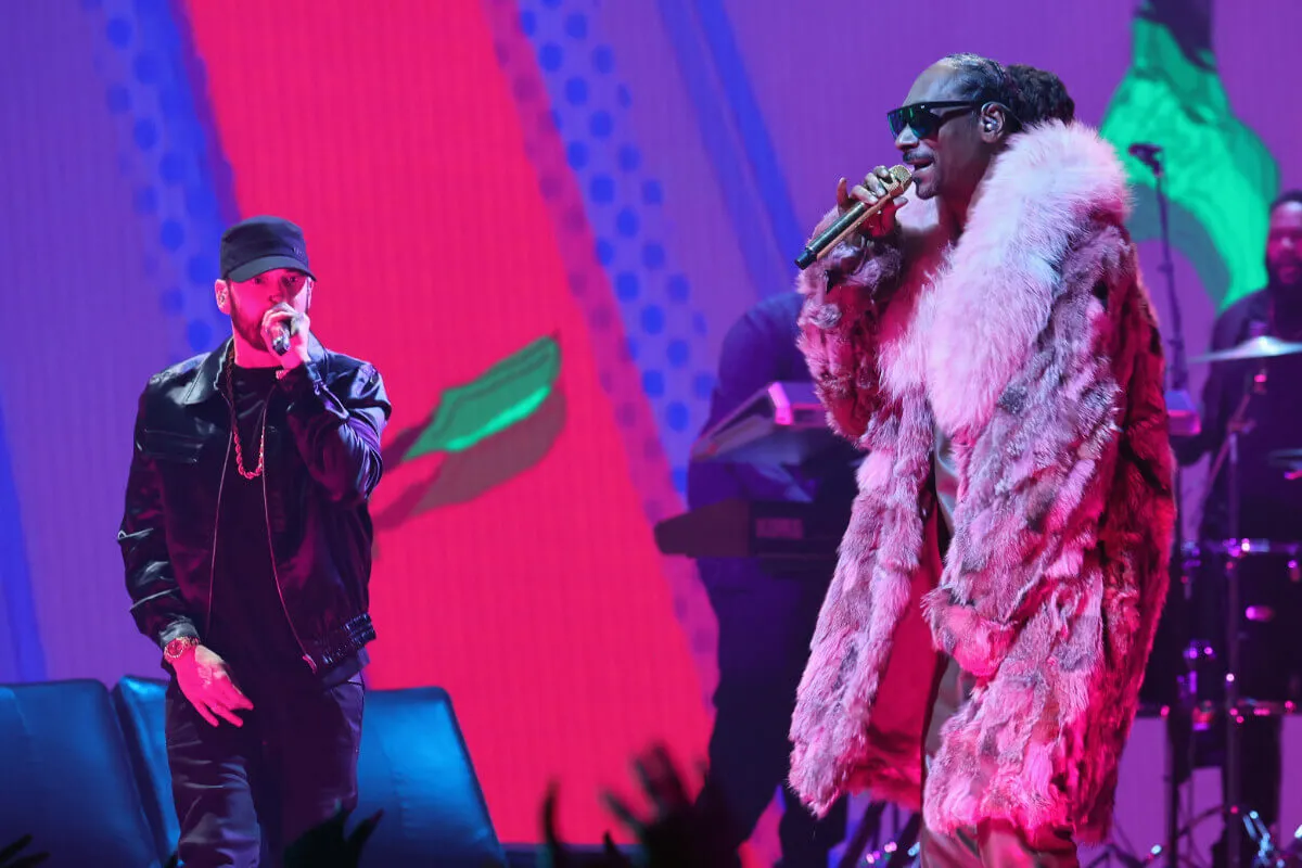 Eminem and Snoop Dogg stand on stage together. They both hold microphones up to their faces. Eminem wears a black jacket and a hat. Snoop wears a coat and sunglasses.