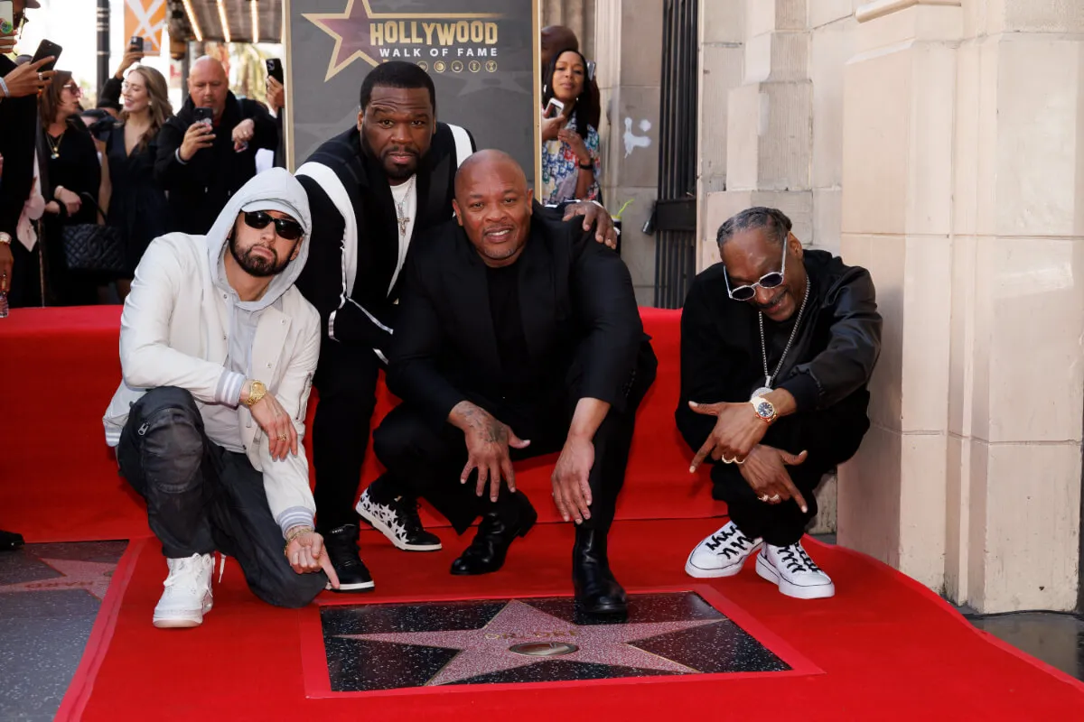 Eminem, 50 Cent, Dr. Dre, and Snoop Dogg crouch over a star on the Hollywood Walk of Fame. Dr. Dre, 50 Cent, and Snoop wear black. Eminem wears white.