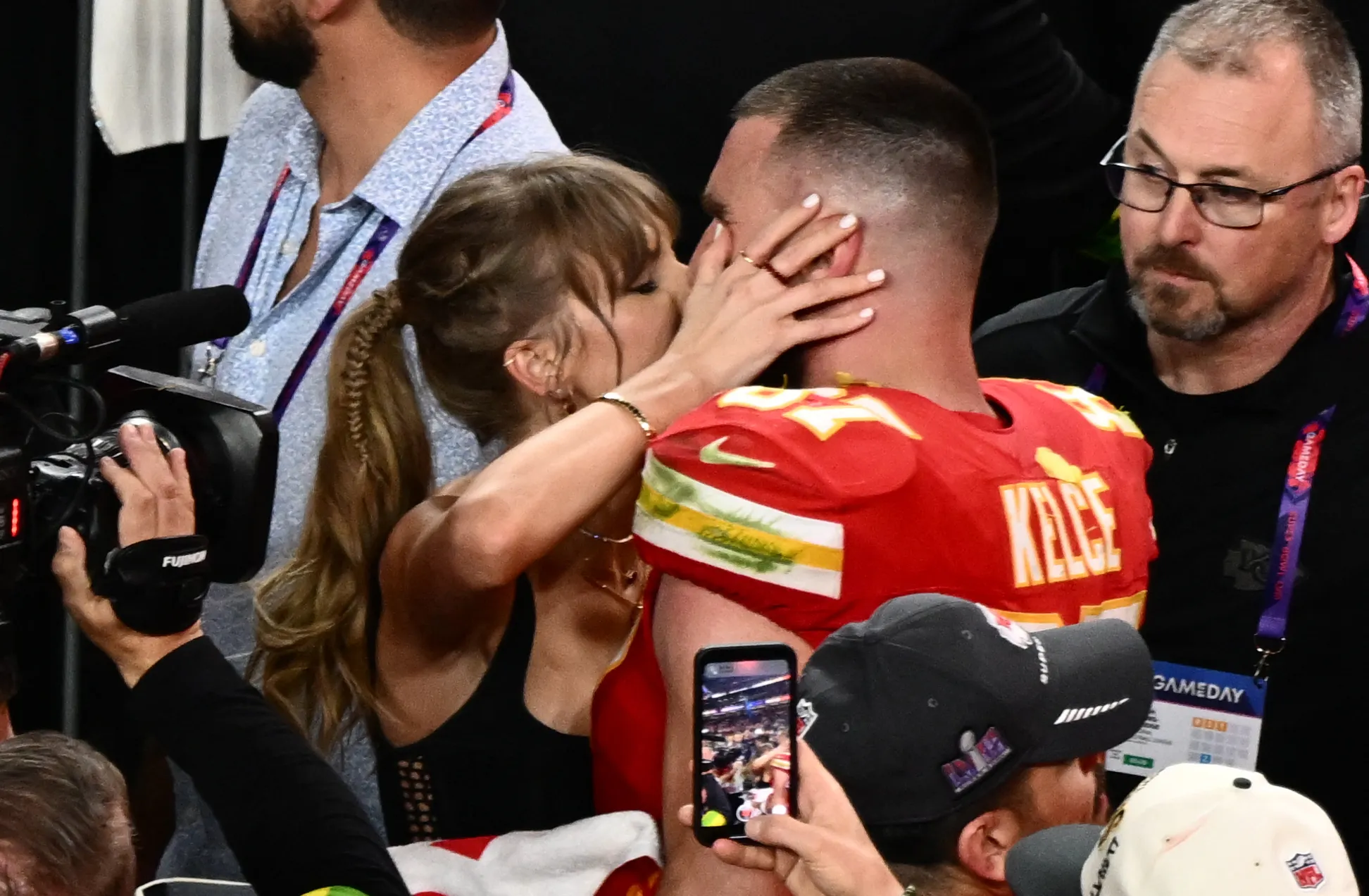 Taylor Swift holding Travis Kelce's head as she kisses him amongst a crowd after the Super Bowl in 2024