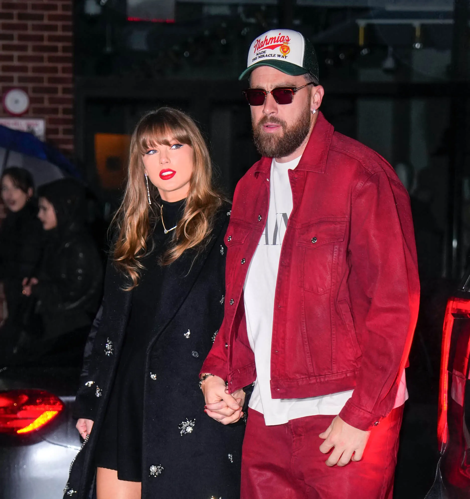 Taylor Swift in a dark dress walking with Travis Kelce at night in New York City, who's dressed in a red jacket and matching pants