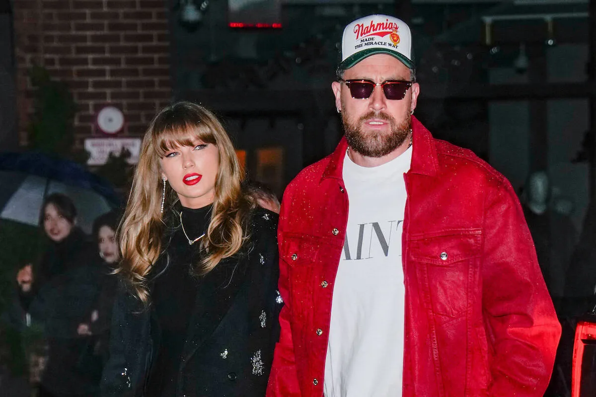 Taylor Swift in a dark dress walking with Travis Kelce, who's in a red jacket and trucker hat, at night in New York City