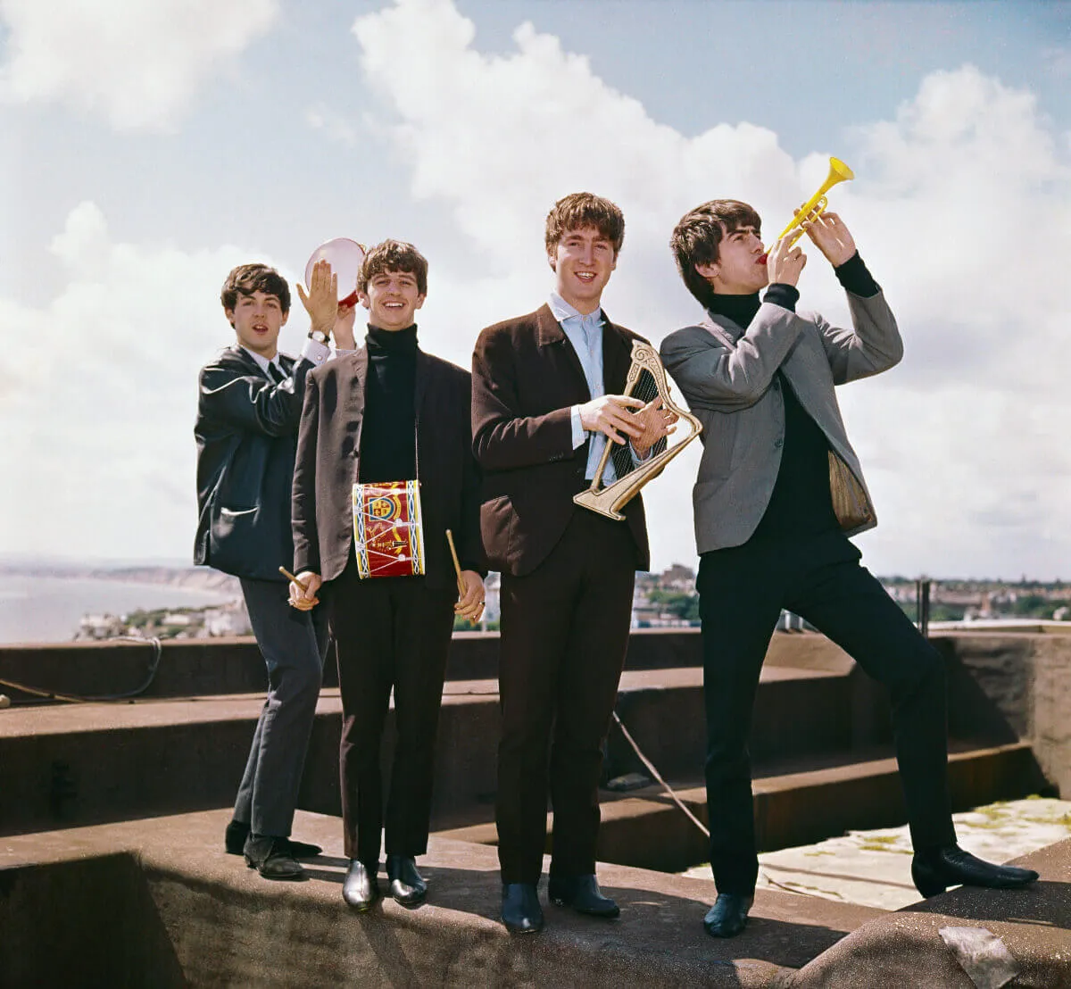 Paul McCartney, Ringo Starr, George Harrison, and John Lennon of The Beatles stand on a rooftop and hold miniature instruments.