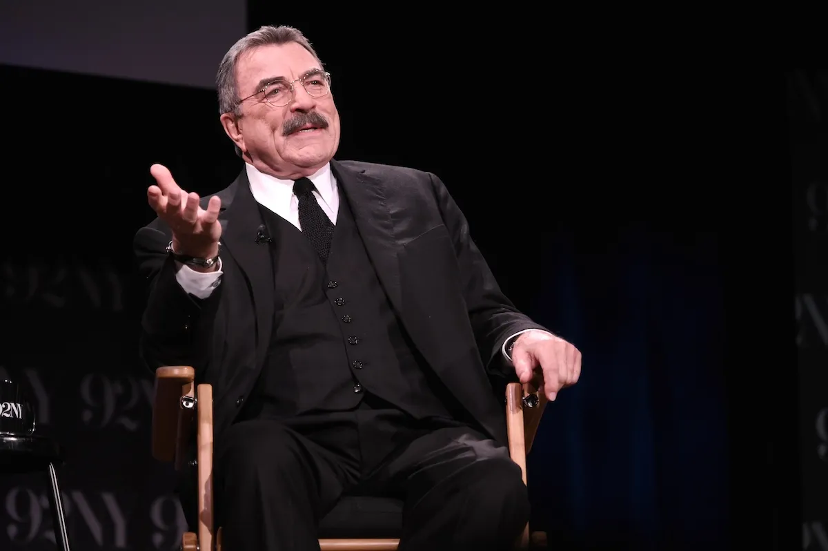 Tom Selleck in a black suit and sitting on stage