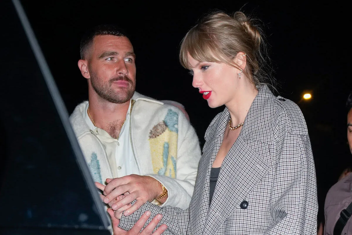 Travis Kelce and Taylor Swift standing next to each other at night. Kelce is in the background and Swift is in the foreground.