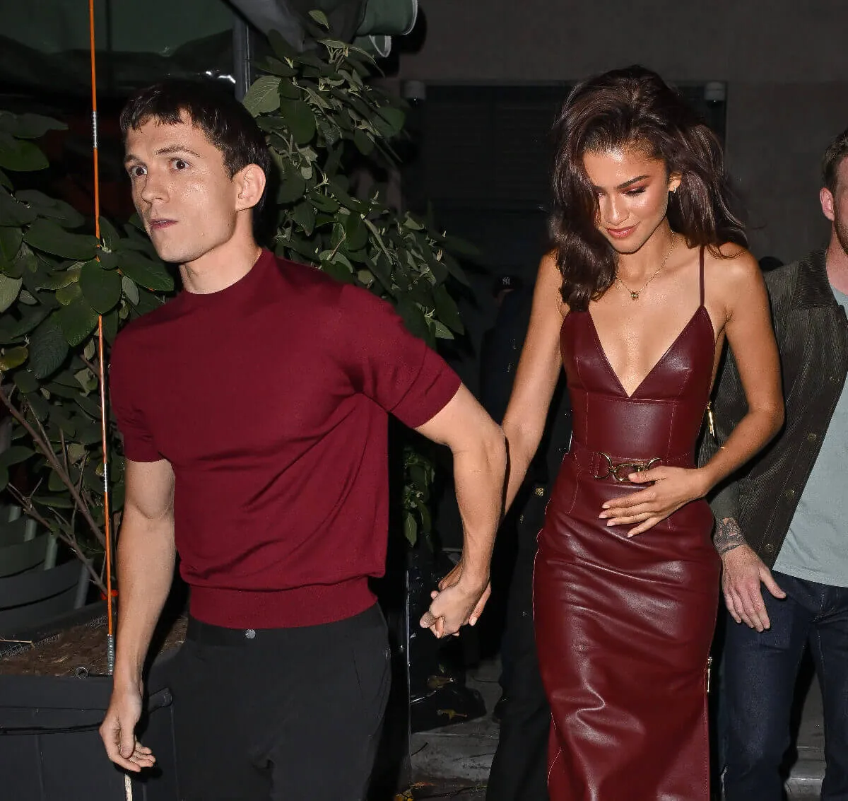 Zendaya and Tom Holland hold hands while walking. They both wear maroon. 