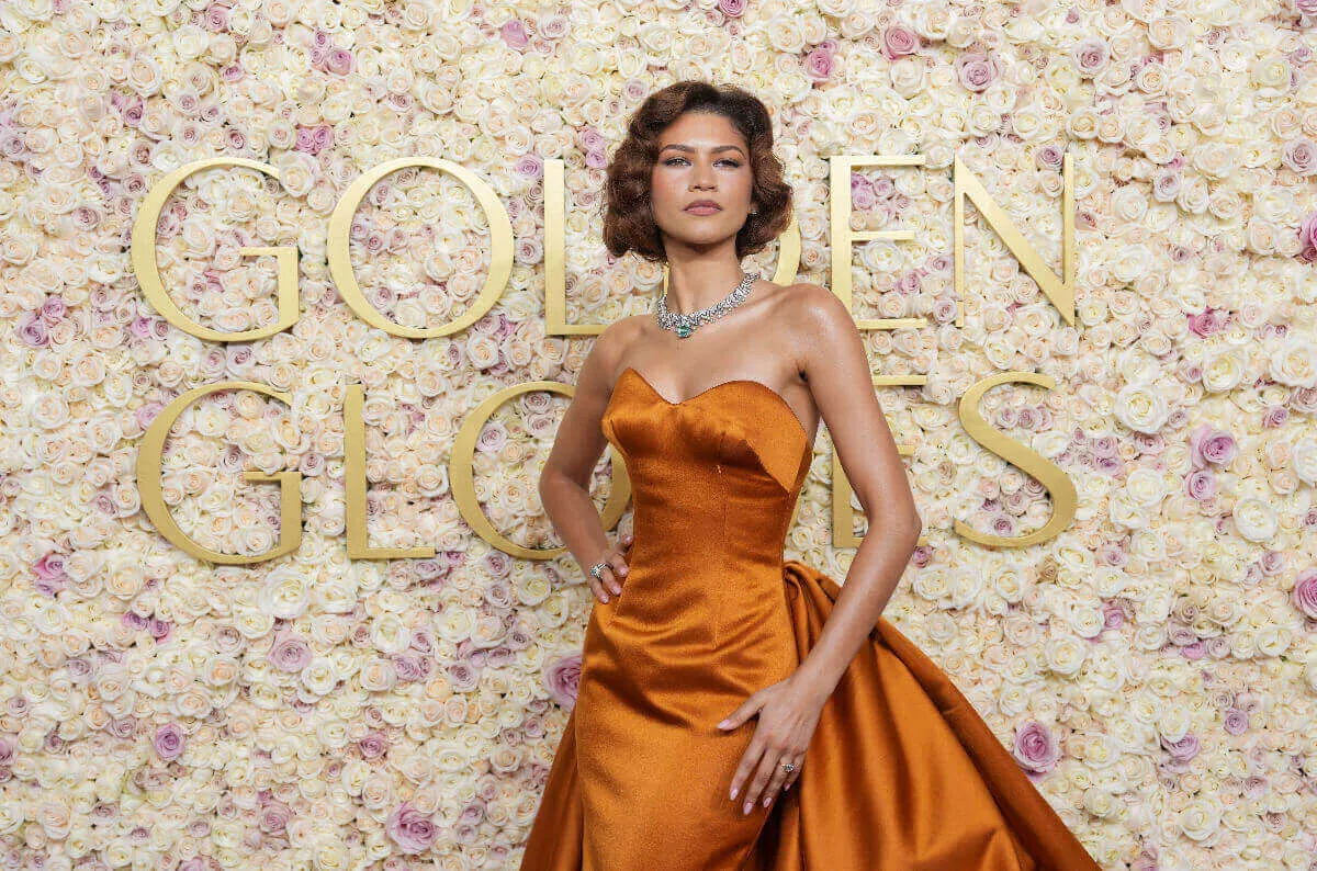 Zendaya wears an orange dress and stands in front of a sign for the Golden Globes.