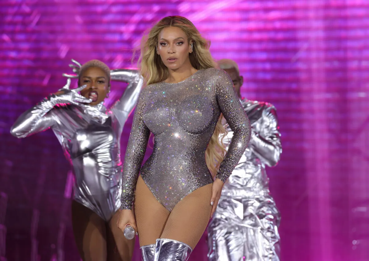 Beyonce wears a silver bodysuit and stands on a stage with two backup dancers behind her.