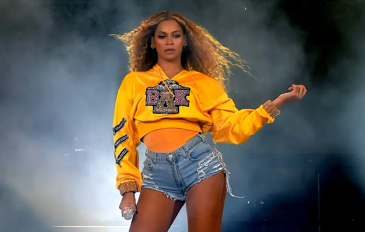 Beyonce wears a yellow sweatshirt and denim shorts. She holds a microphone.