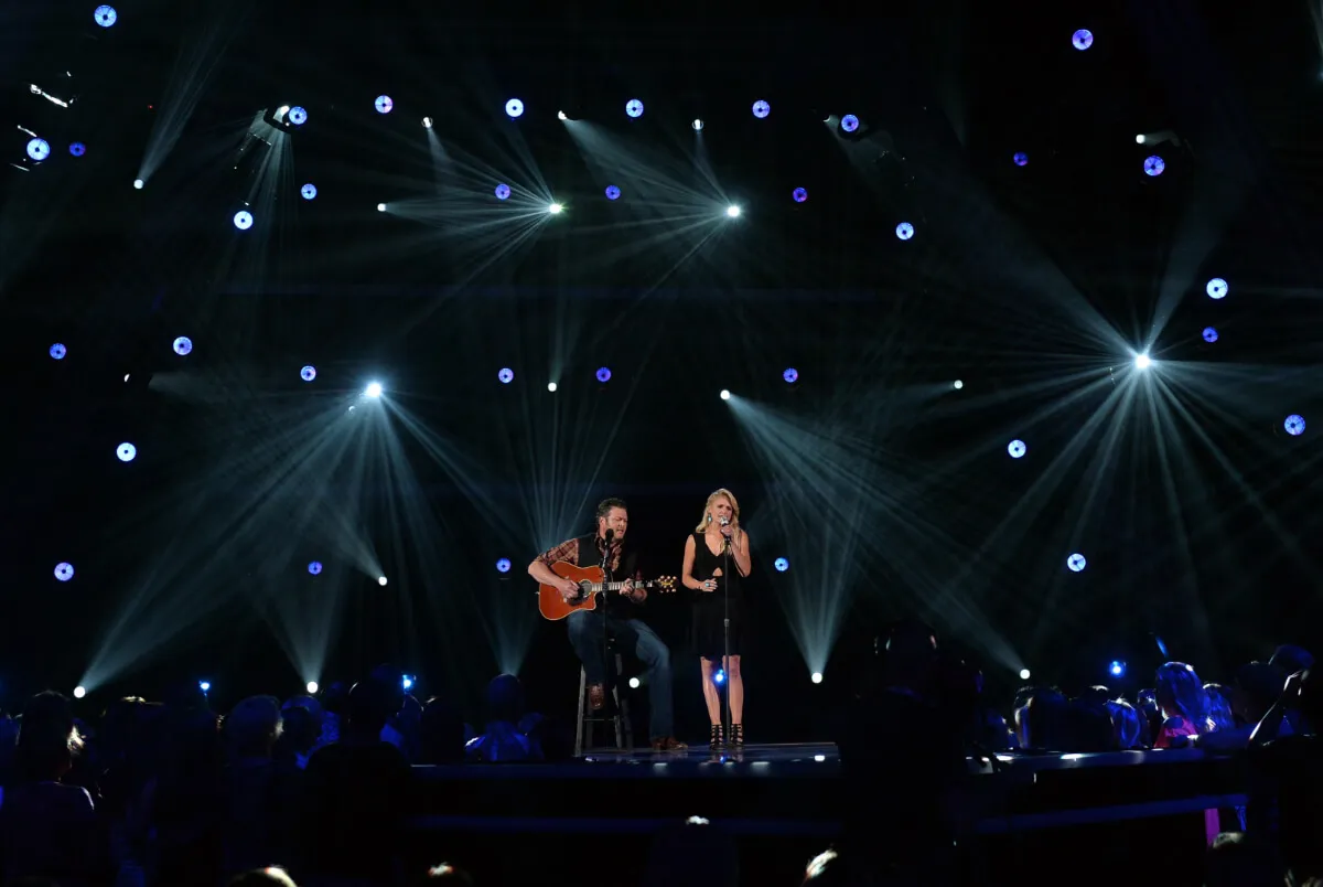 Blake Shelton and Miranda Lambert perform on stage. She stands at the microphone in a black dress and he sits on a stool with a guitar.